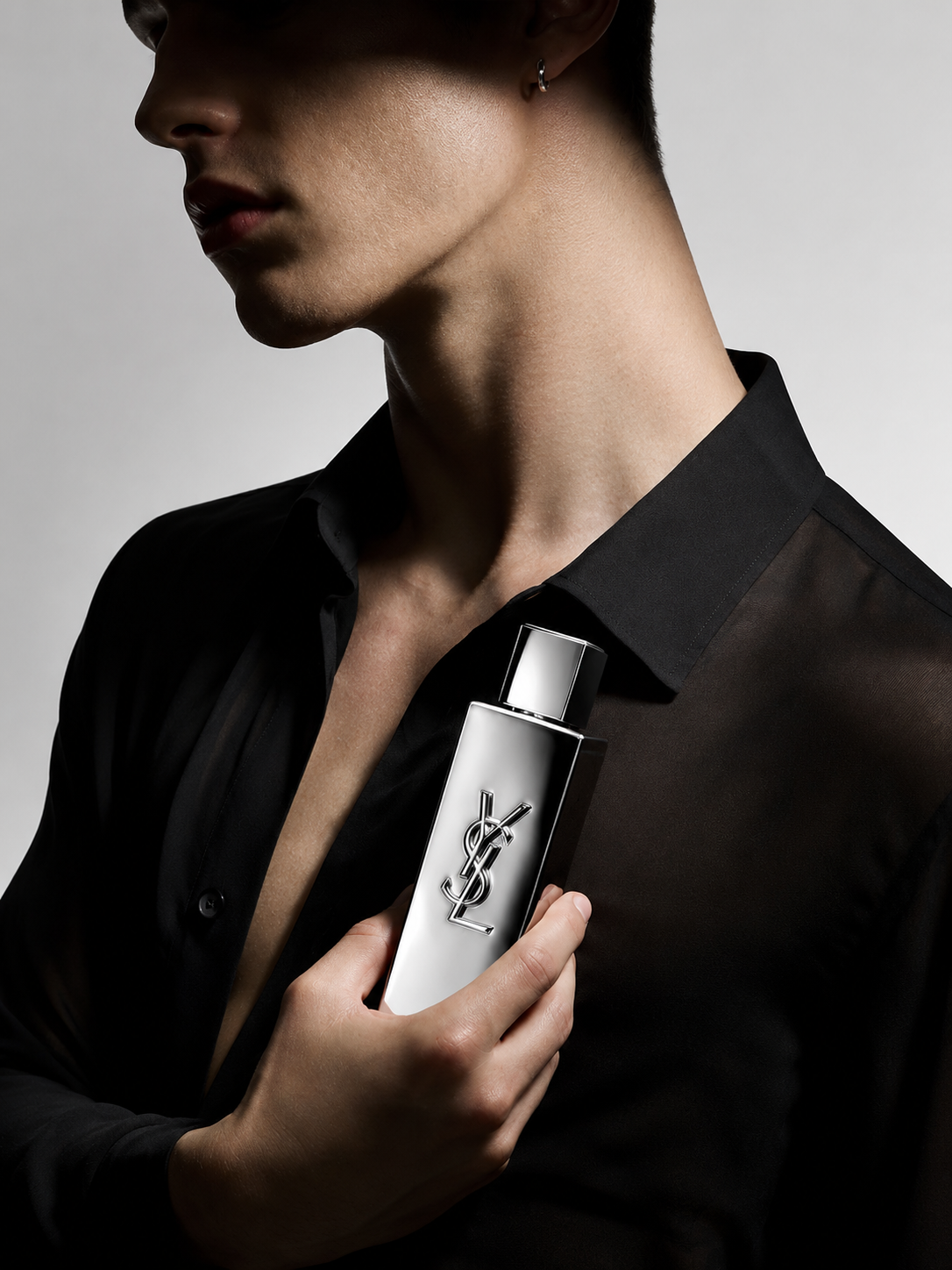 A man in a black shirt holds a silver cylindrical bottle with a YSL monogram logo against a dark gray studio background.