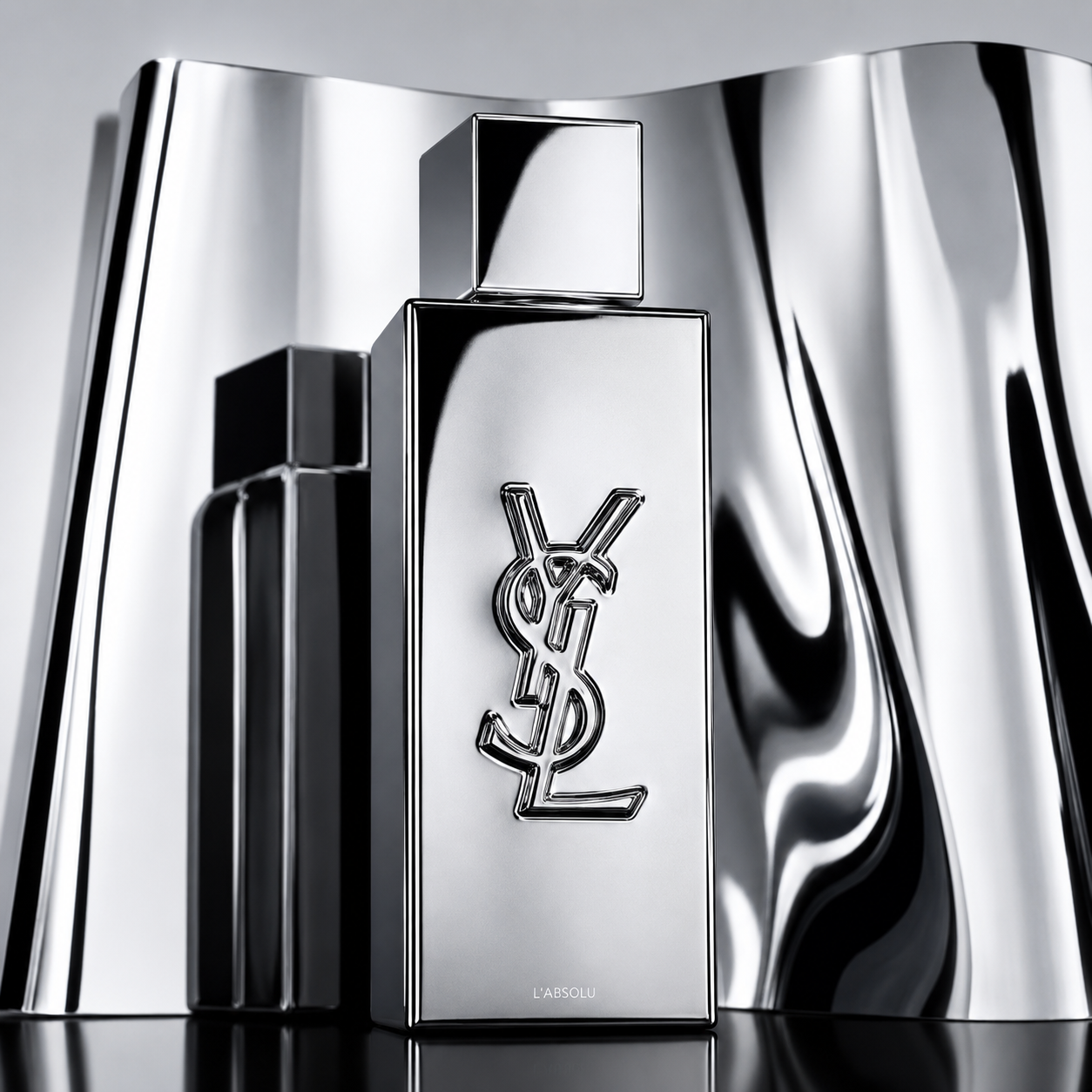 A metallic silver Yves Saint Laurent La Nuit perfume bottle with the interlocking YSL logo, displayed against a reflective, wavy silver background.