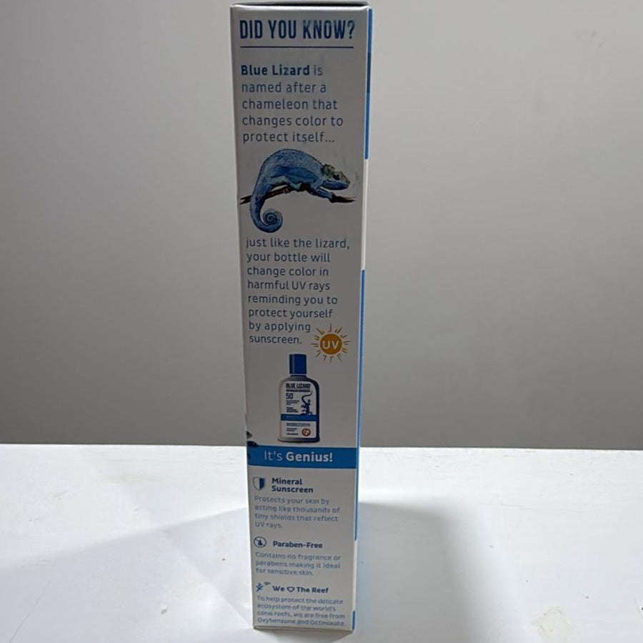 A tall, narrow product box for Blue Lizard brand stands upright on a white surface against a light gray wall. The box is mostly white with blue text and graphics, including a small lizard illustration and a bottle photo near the bottom.
