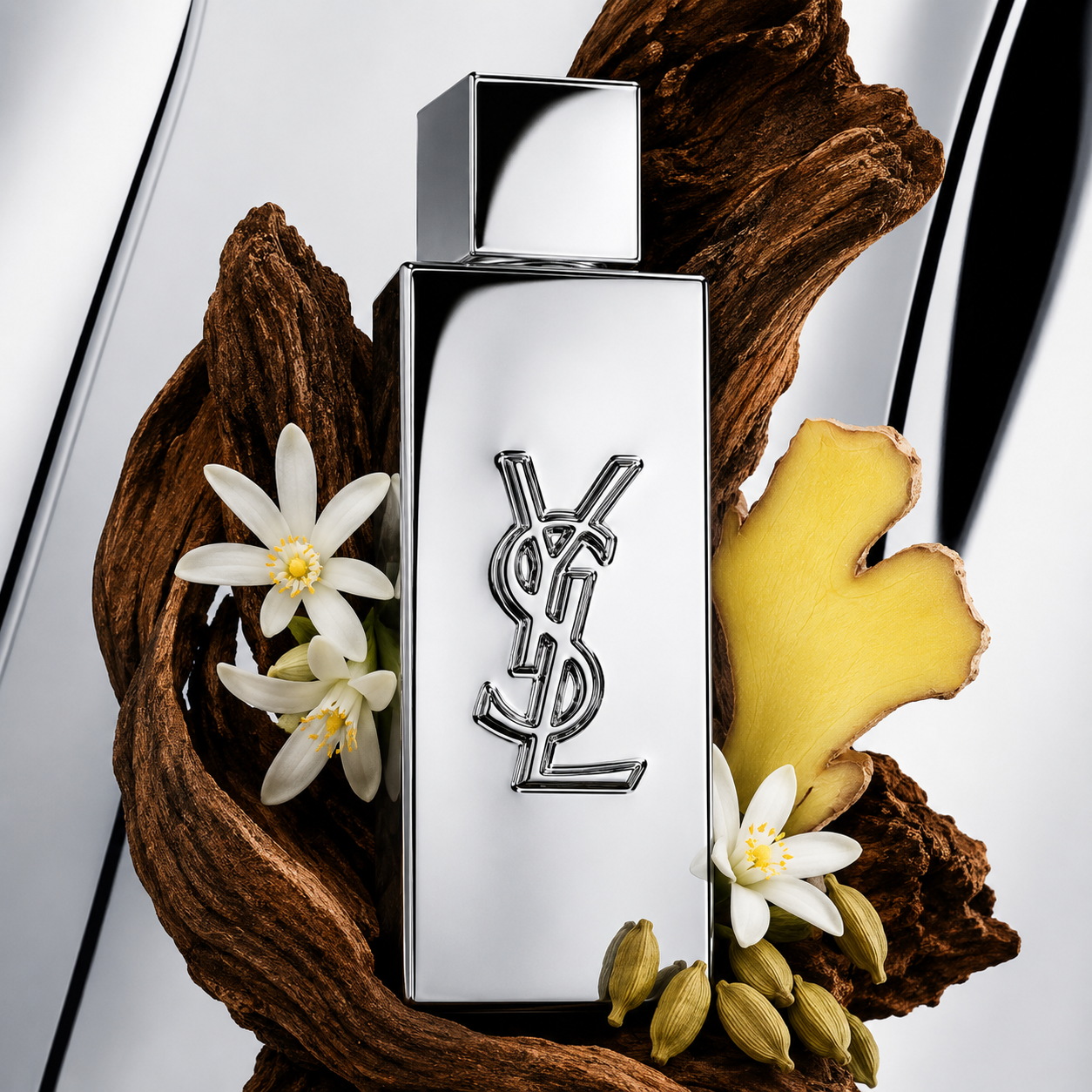 YSL Libre perfume bottle with a reflective silver finish and the iconic YSL logo, styled with white flowers, a yellow ginger root, and green cardamom pods on a dark wood base against a white background.